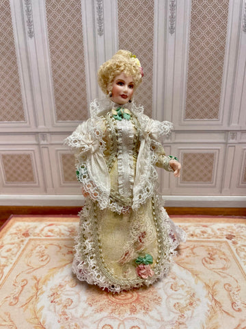 An antique-style 1:12 miniature scale porcelain doll with blonde hair wearing an ornate cream-colored Victorian gown with lace and floral details.