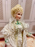 A vintage-style 1:12 miniature scale porcelain doll with curly blonde hair, wearing an ornate cream lace dress and shawl with green floral accents