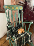 A miniature green rocking chair with yellow floral detailing on the backrest, styled with a plaid blanket and a small orange cat figurine on a wooden floor.
