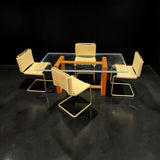 A 1:12 miniature scale modern 5-piece dining set featuring a rectangular glass-top table with a warm orange-brown wooden frame, surrounded by four matching chairs with light tan leather upholstery and gold metal cantilever frames.