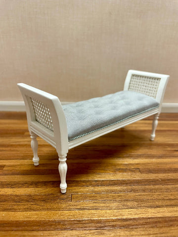 A miniature white wooden settee bench with a tufted light gray cushion and woven cane side rests